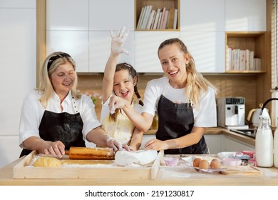 Elderly Granny Rolling Out Dough With Rolling Pin. Young Mother Sprinkle Flour On Dough Happy Little Daughter Raising Hand With Flour Up Showing High Five. Family Cooking Baking In Modern Kitchen.