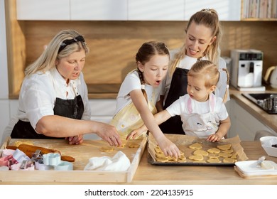 Elderly Granny Teaching Grandkids Putting Cookies On Baking Tray. Surprised Older Daughter Talking To Smaller. Mother Managing Process Watching At. Women Preparing Cooking Apple Pie For Breakfast.