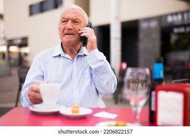 Elderly Gray Haired Man Sitting At Table In Outdoor Cafe And Talking On Phone.