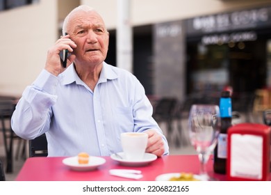 Elderly Gray Haired Man Sitting At Table In Outdoor Cafe And Talking On Phone.