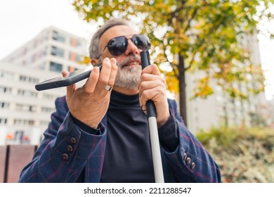 Elderly gray-haired caucasian entrepreneur with low vision in sunglasses sitting on bench, having a phone call using speaker mode, and holding white walking crane with one hand. Outdoor closeup shot