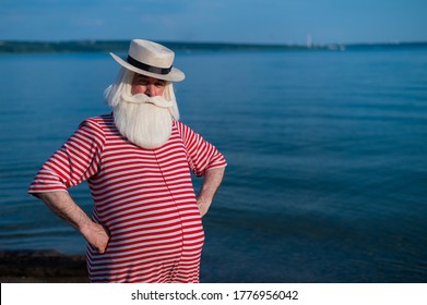 Elderly Gray-haired Man With A Beard In A Striped Bathing Suit And Hat Posing On The Beach. Senior Citizen On Vacation By The Lake.