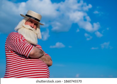 Elderly Gray-haired Man With A Beard In A Striped Bathing Suit And Hat Posing On The Beach. Senior Citizen On Vacation By The Lake.