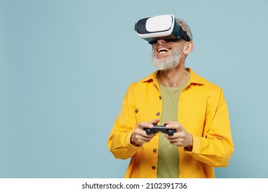 Elderly gray-haired mustache bearded man 50s wear yellow shirt hold in hand play pc game with joystick console watching in vr headset pc gadget isolated on plain pastel light blue background studio.