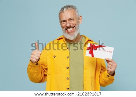 Elderly gray-haired mustache bearded man 50s wear yellow shirt hold store gift certificate coupon voucher card show thumb up isolated on plain pastel light blue background. People lifestyle concept