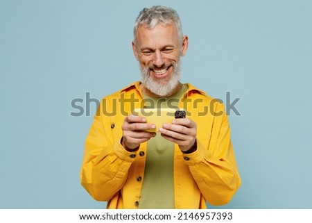 Elderly gray-haired mustache bearded man 50s in yellow shirt using play racing app on mobile cell phone hold gadget smartphone for pc video games isolated on plain pastel light blue background studio.