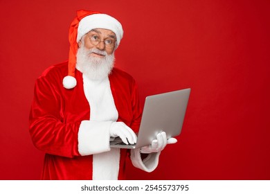 Elderly gray-haired Santa Claus man in Christmas hat red suit coat white gloves glasses working on laptop pc computer isolated on red background. Happy New Year celebration merry holiday concept - Powered by Shutterstock