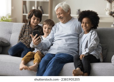 Elderly great-grandfather and three little great-grandchildren having fun with smart phone watch funny videos, enjoy new amusing mobile application at home. Multi-generational people using modern tech - Powered by Shutterstock