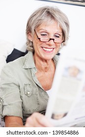 Elderly Grey Haired Lady Wearing A Pair Of Reading Glasses Sitting Reading The Newspaper With A Smile