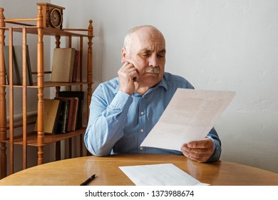 Elderly Grey Haired Man With Mustache Reading A Handheld Document Or Letter With His Hand To His Cheek And A Thoughtful Serious Expression Seated At A Table At Home