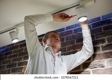 Elderly Grey-haired Man Repairs Lamp Under Ceiling.