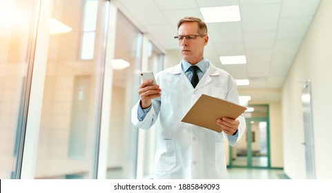 An elderly handsome serious doctor in a white coat and tie walks along the corridor of the clinic holding a patient card or documents in his hand and writes a message on a smartphone. - Powered by Shutterstock