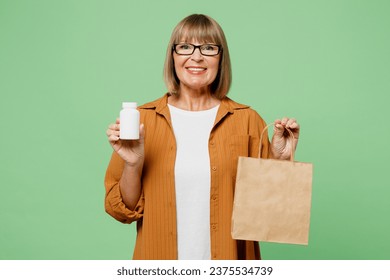 Elderly happy fun woman 50s year old wear brown casual clothes glasses hold in hand bottle of pills blank craft paper packet bag isolated on plain green background studio. Delivery service from shop - Powered by Shutterstock