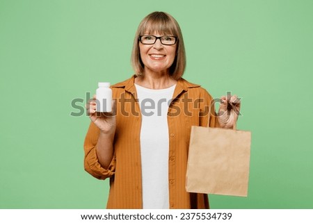 Elderly happy fun woman 50s year old wear brown casual clothes glasses hold in hand bottle of pills blank craft paper packet bag isolated on plain green background studio. Delivery service from shop