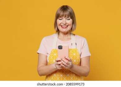 Elderly Happy Housekeeper Housewife Woman 50s In Orange Apron Hold In Hand Mobile Phone Search Look For Recipe Isolated Plain On Yellow Background Studio Portrait. People Household Lifestyle Concept