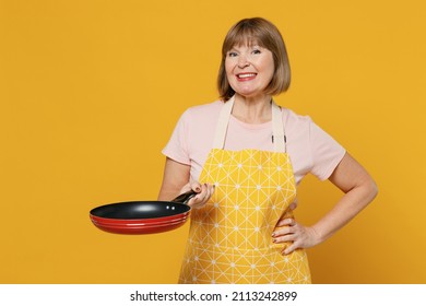 Elderly Happy Smiling Housekeeper Housewife Woman 50s In Orange Apron Hold Empty Frying Pan Cooking Stand Akimbo Arm On Waist Isolated Plain On Yellow Background. People Household Lifestyle Concept