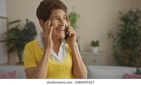 Elderly hispanic woman with short hair talking on the phone indoors, smiling while sitting in a living room filled with plants and home decor. - Powered by Shutterstock