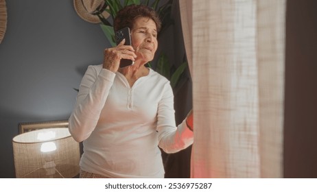 Elderly hispanic woman with short hair standing indoors in her living room and talking on a smartphone while looking out the window at the reflected light from a police car. - Powered by Shutterstock