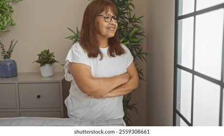 An elderly hispanic woman stands indoors with arms crossed, gazing out the window in her living room, surrounded by plants, reflecting a calm and thoughtful atmosphere. - Powered by Shutterstock