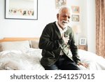 Elderly Indian man sitting on bed, clutching chest in discomfort. Senior male experiencing chest pain. Concerned elderly man in bedroom setting. Elderly Indian man unwell with chest pains.