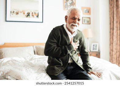 Elderly Indian man sitting on bed, clutching chest in discomfort. Senior male experiencing chest pain. Concerned elderly man in bedroom setting. Elderly Indian man unwell with chest pains. - Powered by Shutterstock