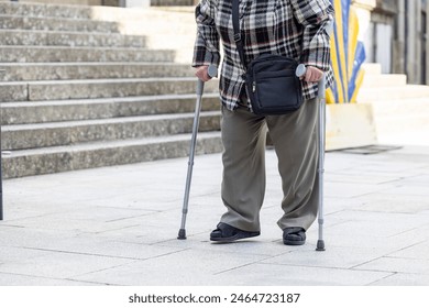 An elderly individual is seen from the waist down utilizing two walking canes for mobility assistance on a city street - Powered by Shutterstock