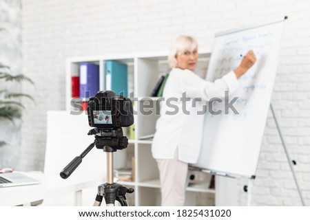 Elderly intelligent woman is writing on flipchart and recorded video classes on the camera, side view. Online teacher, video learning concept