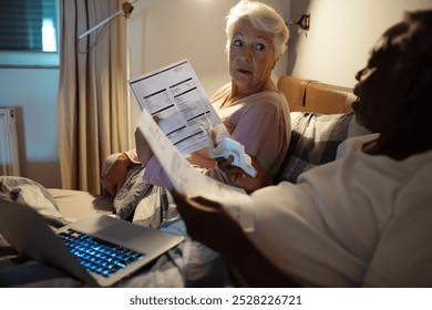 Elderly interracial couple managing finances in bed with laptop - Powered by Shutterstock