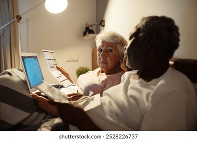 Elderly interracial couple managing finances in bed with laptop - Powered by Shutterstock