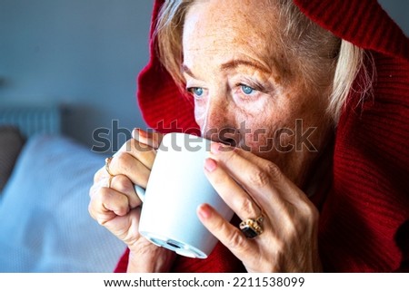 elderly lady drinking hot tea while freezing at home due to lack of heating; pensioners freezing at home; energy crisis 2022; cold at home;  European energy crisis, blackout in Europe