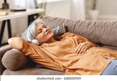 Elderly lady in headphones lying on couch and listening to music with closed eyes while resting in cozy living room at home