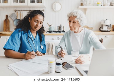 Elderly lady sitting at kitchen table near african american nurse in blue uniform writing prescriptions after giving her first aid, senior woman looking attentively at notes, holding bottle of water - Powered by Shutterstock