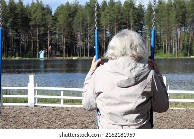 An Elderly Lady Is Sitting On A Swing By A Lake On A Sunny Day. She Is Thinking About The Past. Concepts Of Old Age, Memories And Time. 