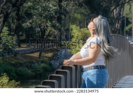 Elderly Latin American tourist woman in profile standing on a bridge looking up with green trees in blurred background, enjoying a walk in a public park, long gray hair, sunglasses, white blouse
