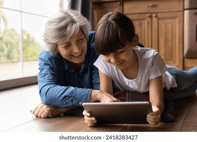 Elderly latina grandmother rest on warm floor with preteen grandkid touch tablet screen in girl hands explain learning material in playful way. Little girl play online game with loving granny on pad - Powered by Shutterstock