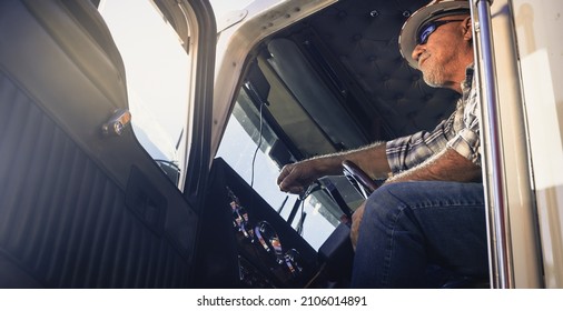 Elderly Latino Semi Truck Driver Sits In The Cab Looking Ahead Wearing Plaid Shirt, Sunglasses And Hard Hat. Handsome Cool Looking Trucker Sits At The Steering Wheel.  