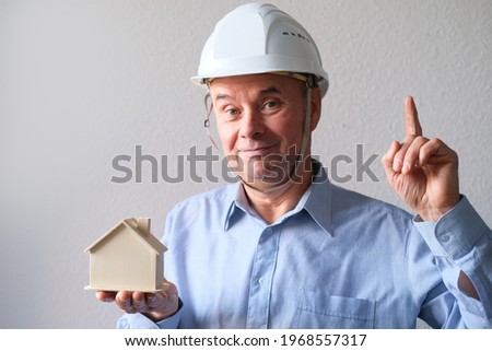 elderly male agent, a builder in a protective helmet, glasses offers to rent, buy a house, shows a model of a house, a mortgage concept, advice on buying a property