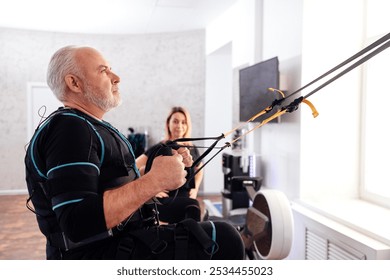 Elderly male athletic in electrical muscle stimulation suit uses trx system for his workout with personal trainer. Greyhaired man exercising with suspension training equipment hanging at fitness club. - Powered by Shutterstock