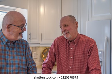 An Elderly Male Gay Couple In Their Kitchen Smiling At Each Other While Having A Conversation.