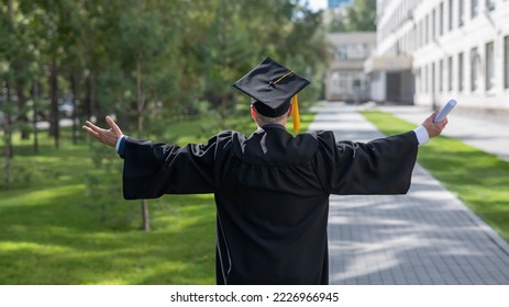 Elderly male graduate rejoices at graduation outdoors. Rear view of a man in a robe with arms outstretched to the sides. 
