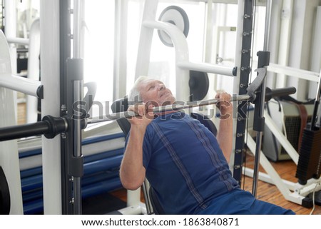 Elderly male is having intense workout in gym. Senior caucasian man lifting weights at fitness club. Strength workout for seniors.