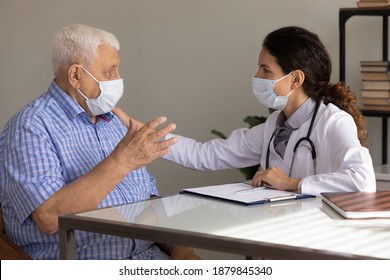 Elderly Male Patient In Medical Facemask Talk Consult At Female Doctor Office In Private Clinic. Caring Woman GP Have Consultation With Senior Man, Wear Facial Masks Against Covid-19 Corona Pandemics.