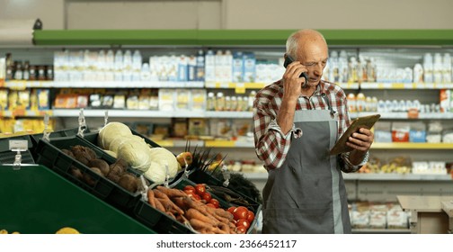 Elderly male sales assistant in a grocery using digital tablet and talking on cell phone while taking inventory. Banner. Copy space - Powered by Shutterstock