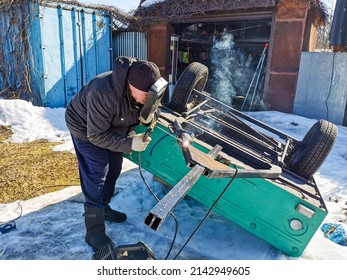 An elderly male welder repairs a car trailer with electric arc welding on a spring sunny day.