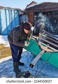 An elderly male welder repairs a car trailer with electric arc welding on a spring sunny day.