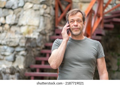 An Elderly Man 45-50 Years Old Is Talking On A Mobile Phone Against A Neutral Background Of The Old City.