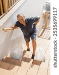 Elderly man with ache expression, leans forward, holds his back because of back pain after climbing the stairs carrying groceries holding onto railing. Twisted face. Vertical