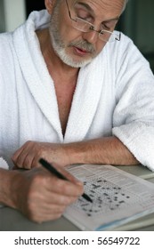 Elderly Man In Bathrobe Sitting At A Table Doing Crosswords