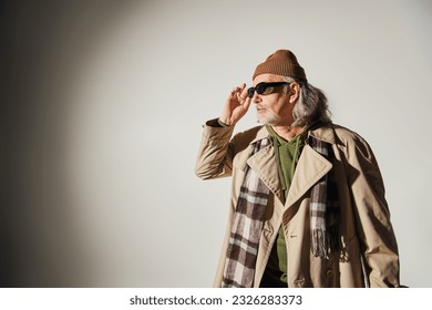 elderly man in beanie hat, beige trench coat and plaid scarf adjusting dark sunglasses and looking away on grey background, hipster style, individuality, fashion and age concept - Powered by Shutterstock