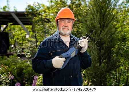 A elderly man with a beard in a hardhat and glasses with a reciprocating saber saw. construction work. compliance with safety regulations. employment in retirement.